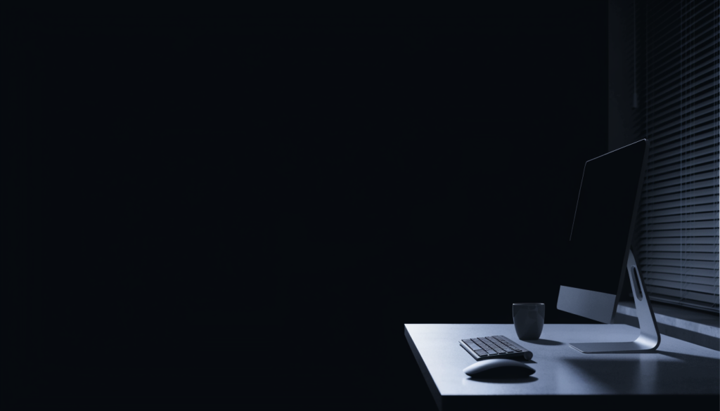 Minimal workspace at night with blank laptop screen and cursor, illuminated by soft desk lamp, suggesting the moment when creative decisions freeze.